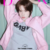 TWS's Kyungmin wearing a pink jacket over a gray shirt with text, sitting on a light blue couch.