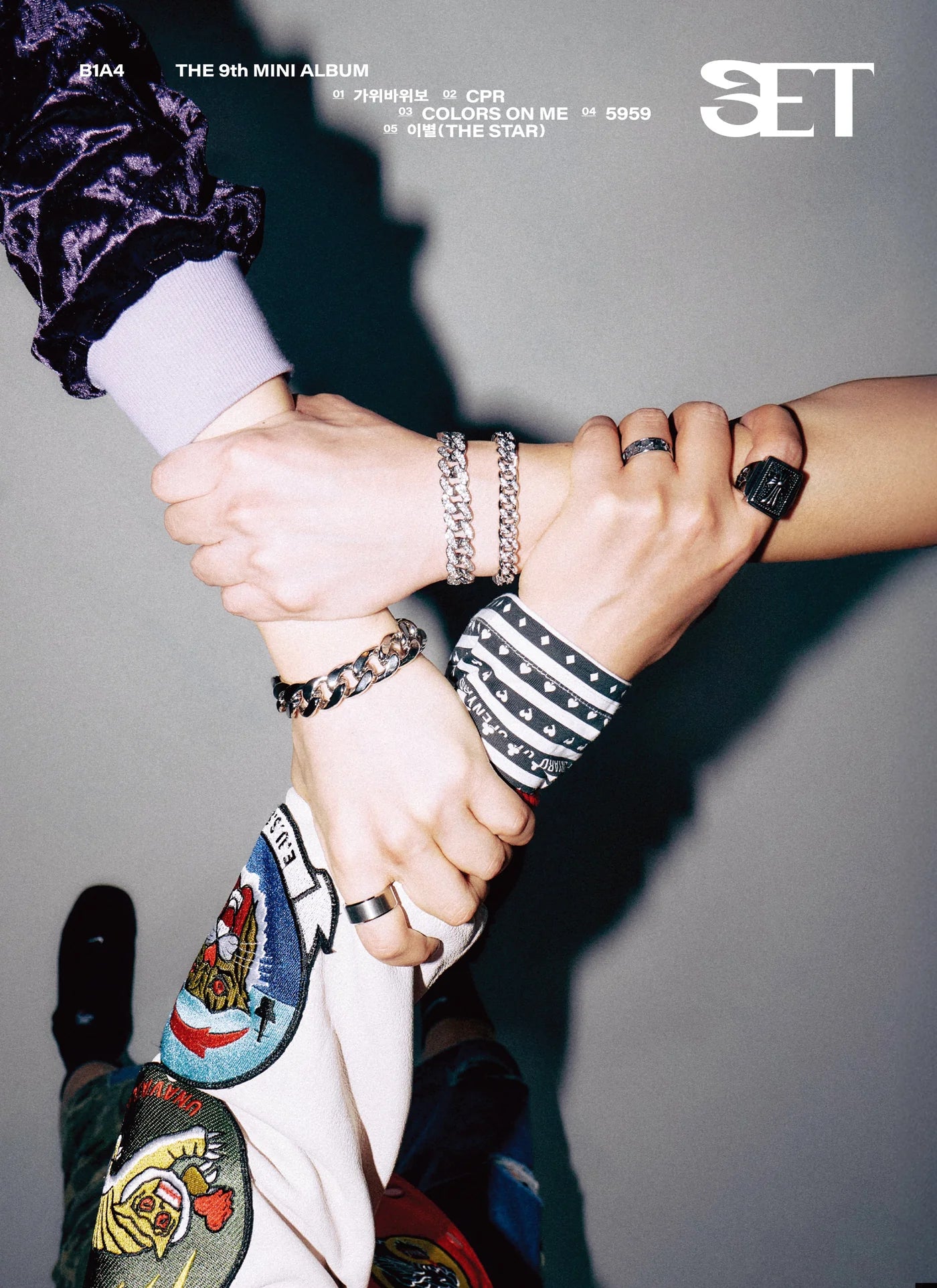 Three people - B1A4 members -  holding hands with visible jewelry against a gray background